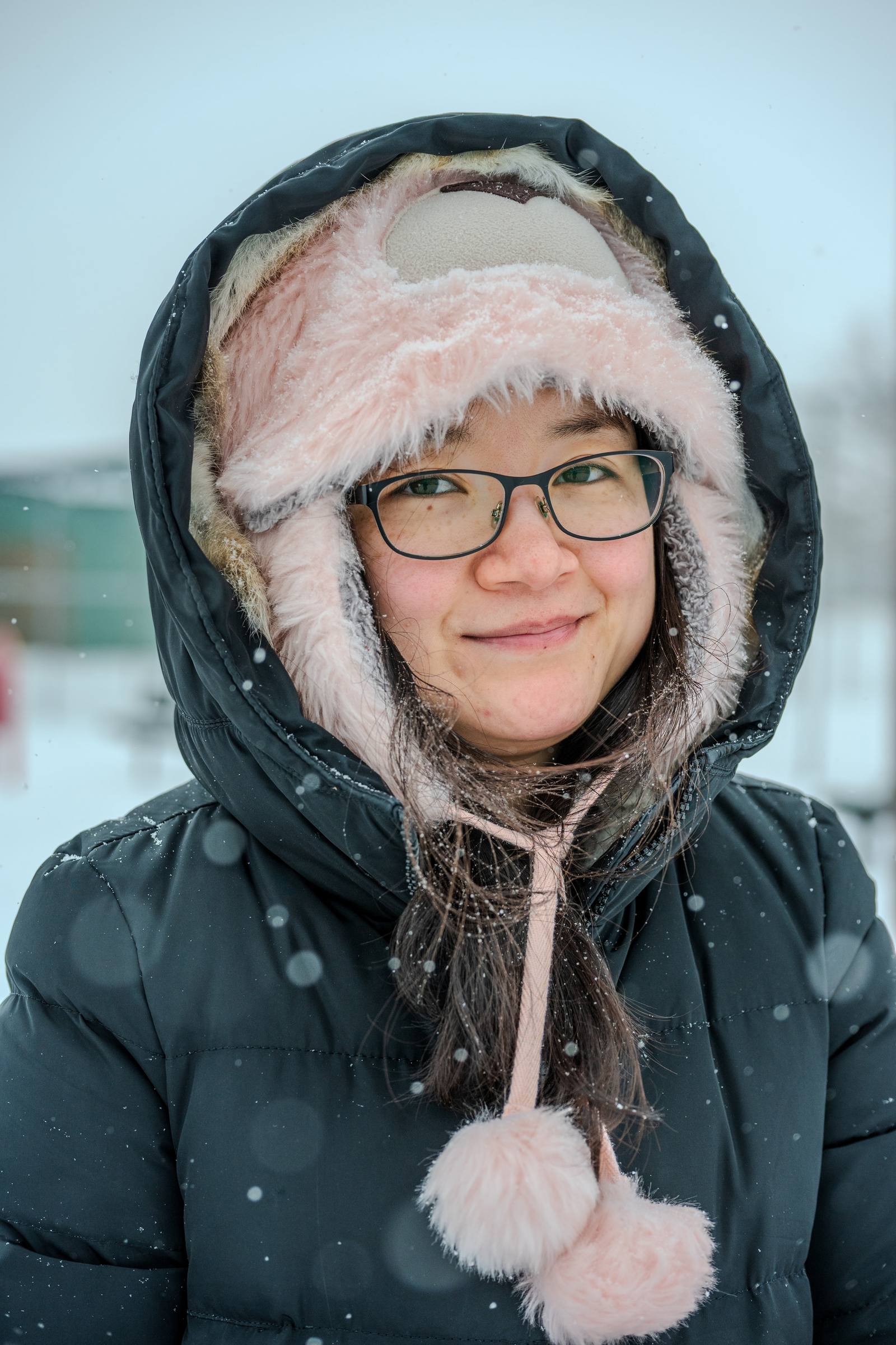 A smiling person in a winter hat and glasses outdoors in falling snow.