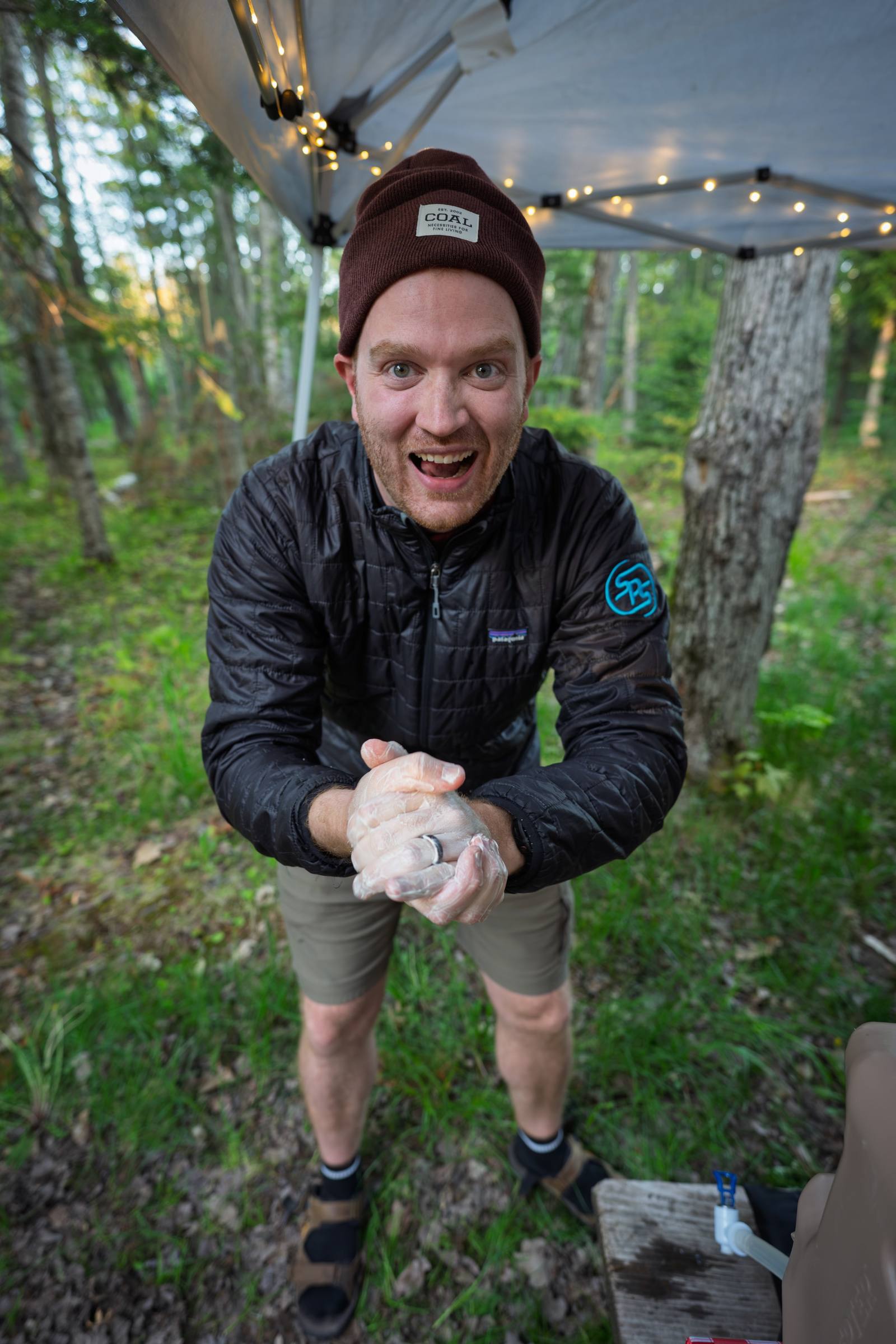 A grinning person in a dark jacket rubs their hands together under a string-lit canopy shelter at a wooded campsite.