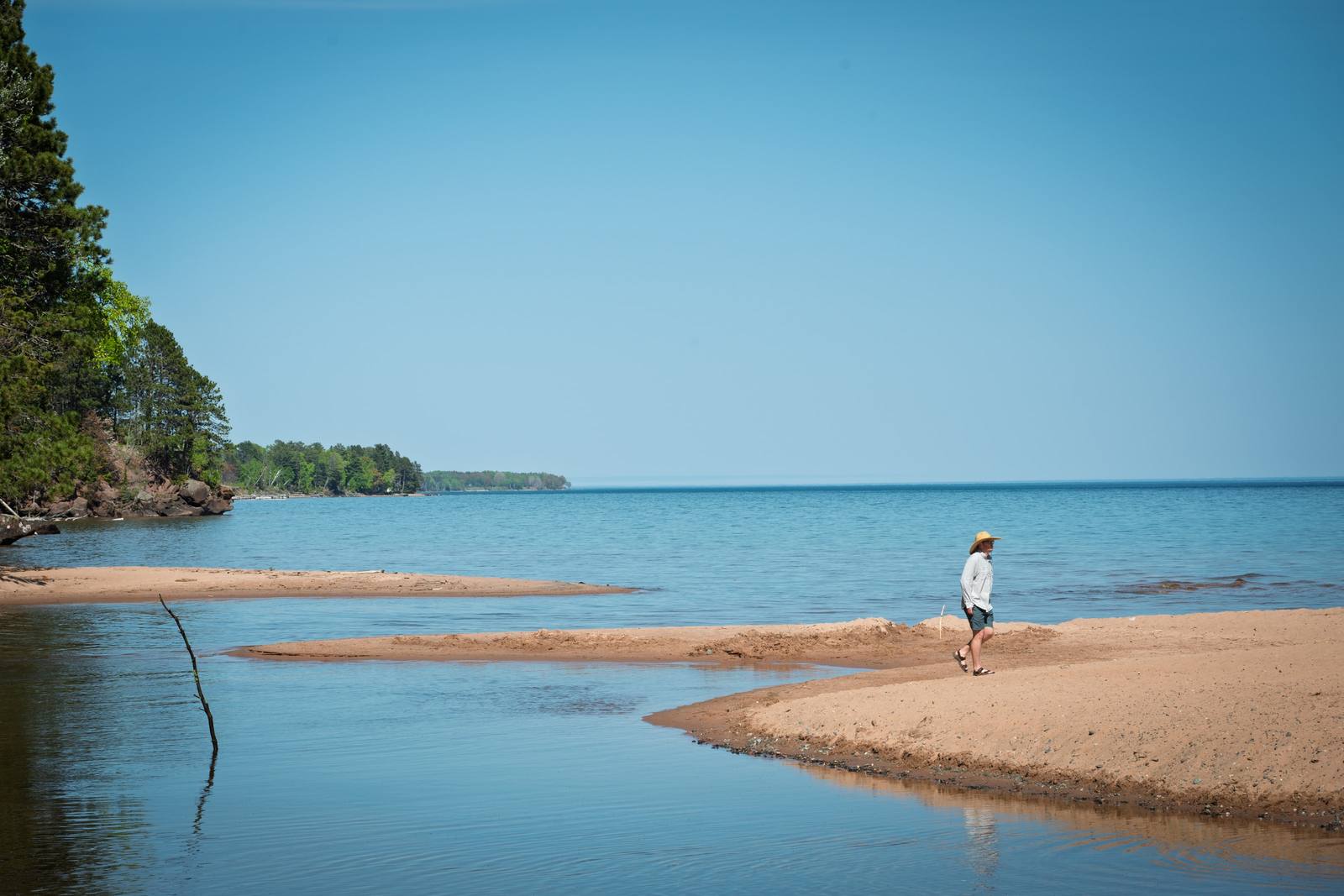 A person in a sun hat strolls along a narrow sandy spit where a small river meets the vast blue waters of a large lake, with a pine-forested rocky shoreline curving away to the left.