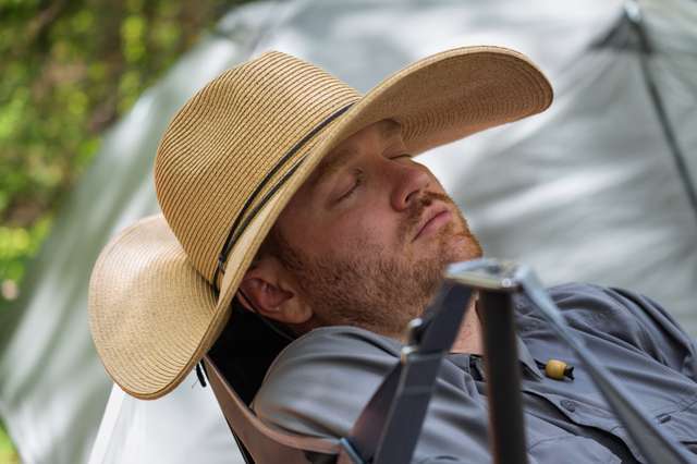 A bearded man in a wide-brimmed straw hat reclines in a camping chair with eyes closed beside a tent in the woods.