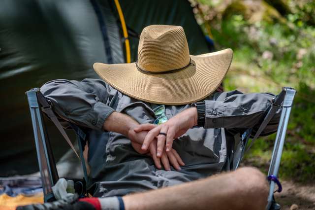 A camper in a wide-brimmed straw hat naps with hands folded in a camping chair beside a tent at a lakeshore campsite.