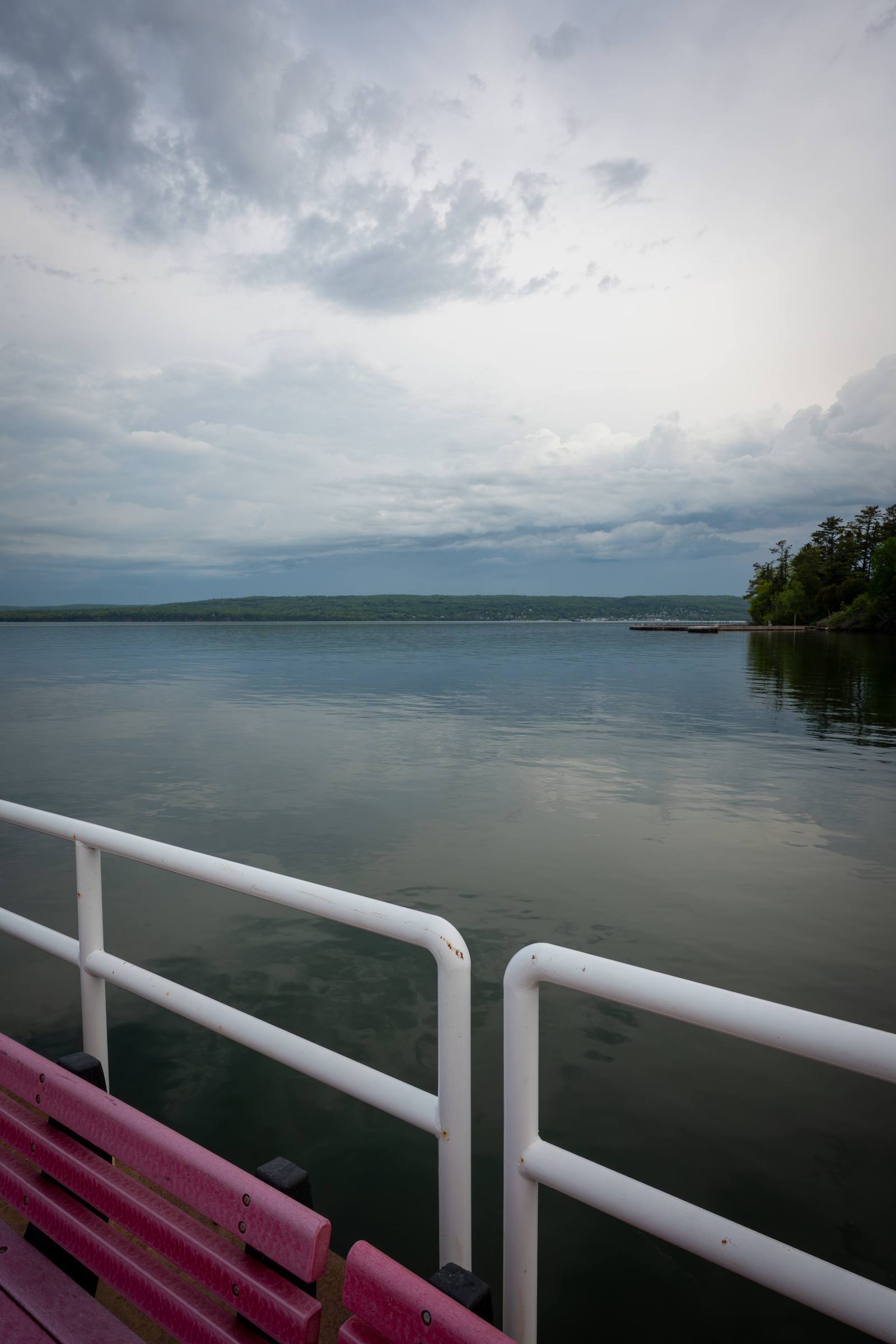 A pink bench on a dock with white railings looks out over a calm lake under a stormy sky, with a forested island on the right and green hills across the water.