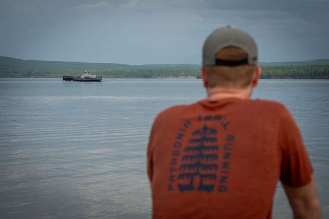 A person in an orange shirt stands at a lakeshore and watches a ferry cross toward a green forested hillside under an overcast sky.