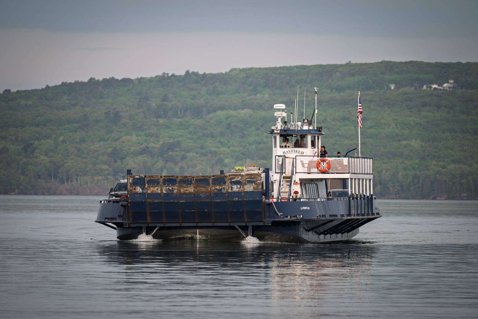 The Bayfield ferry crosses a calm grey lake carrying vehicles toward the forested shore, with an American flag flying from its wheelhouse.