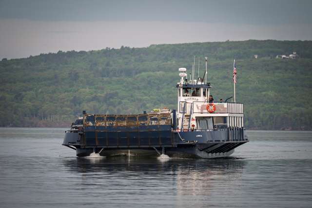 The Bayfield ferry crosses a calm grey lake carrying vehicles toward the forested shore, with an American flag flying from its wheelhouse.