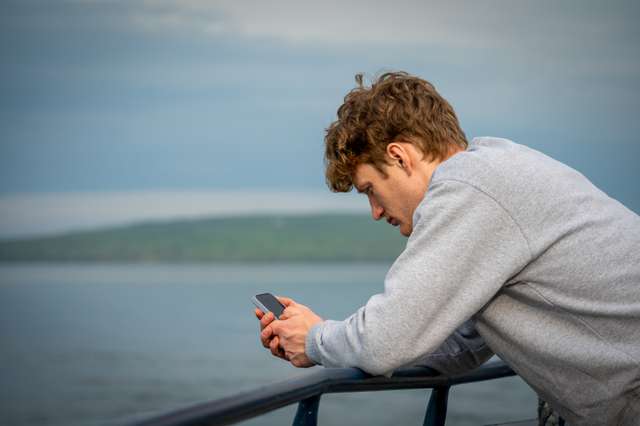 A young man in a grey sweatshirt leans on a ferry railing and looks at his phone, with a green-forested island visible across calm water in the background.