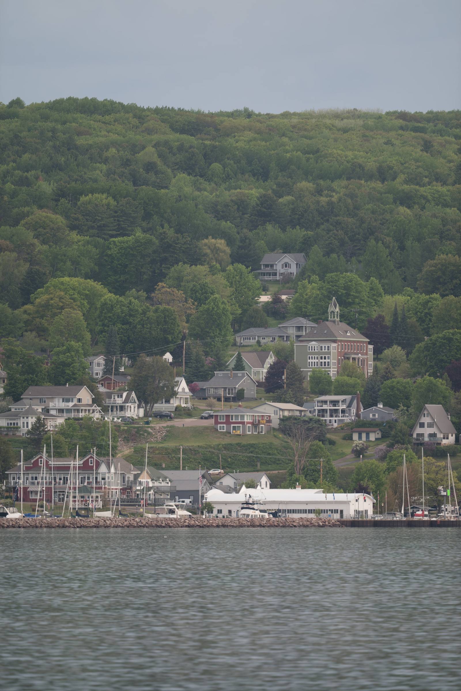 A lakeside town viewed from the water, with a marina full of sailboat masts, colorful storefronts, and a historic brick Victorian building set against a forested hillside.